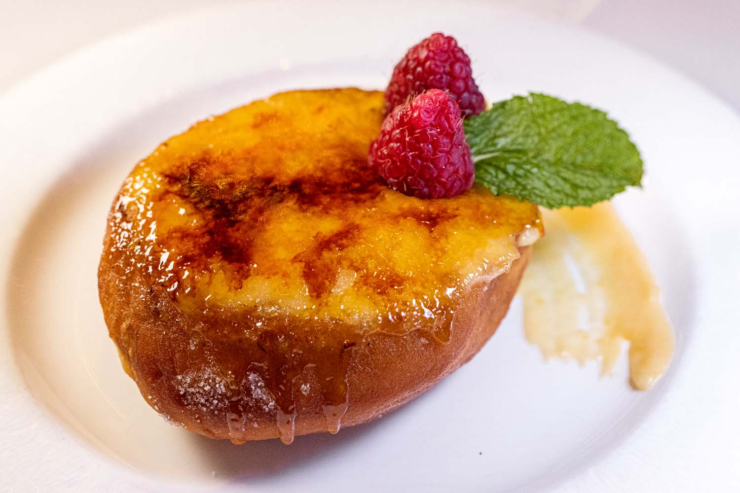 A caramelized donut filled with zabaglione cream and flambéed with peach butter, topped with fresh raspberries and mint leaf on a white plate