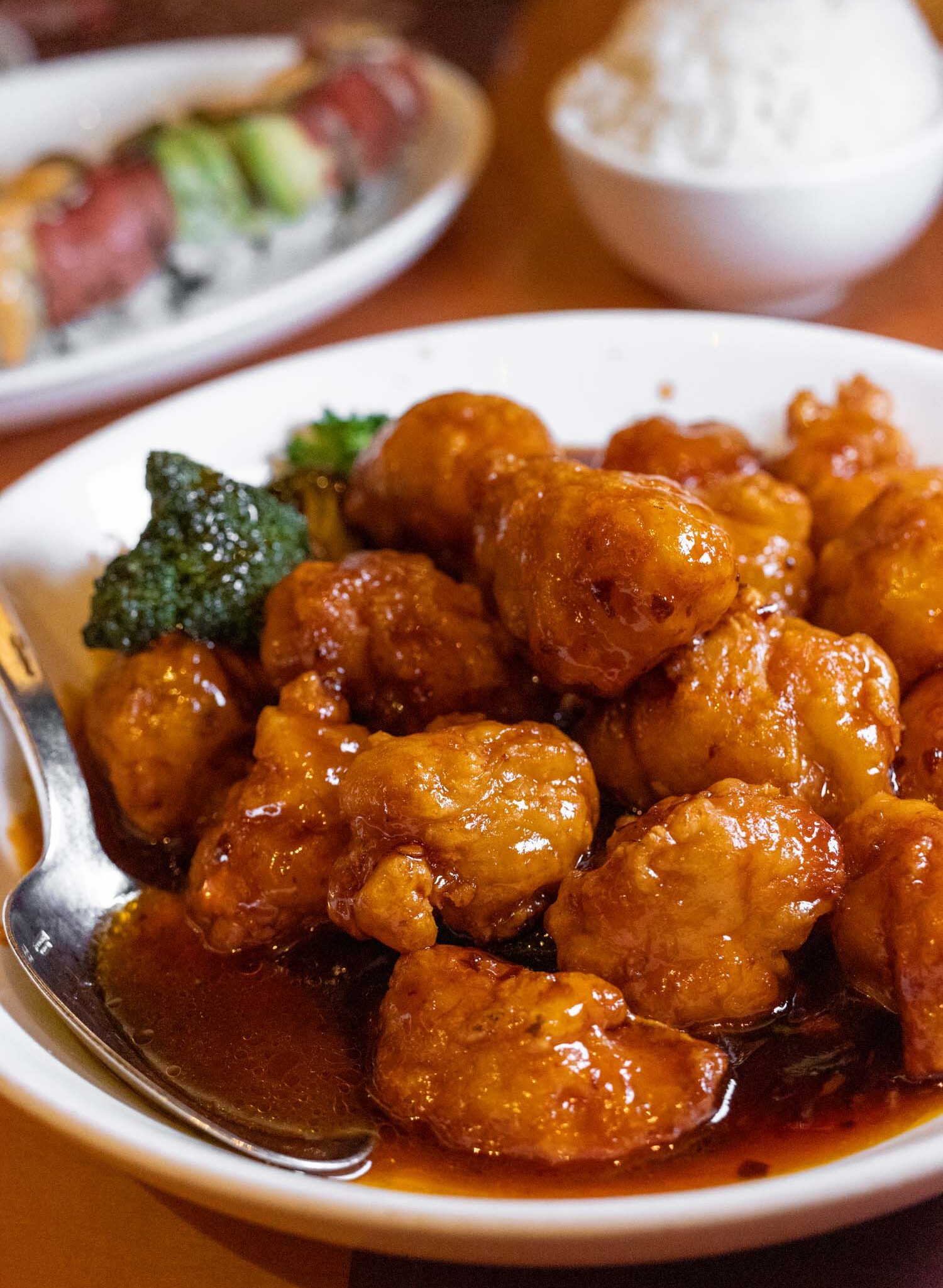 Wok-fried General Tso's chicken glazed in glossy sweet-spicy sauce, garnished with fresh steamed broccoli florets, served in a white bowl with sushi visible in soft focus background, showcasing the dish's characteristic deep golden-brown color