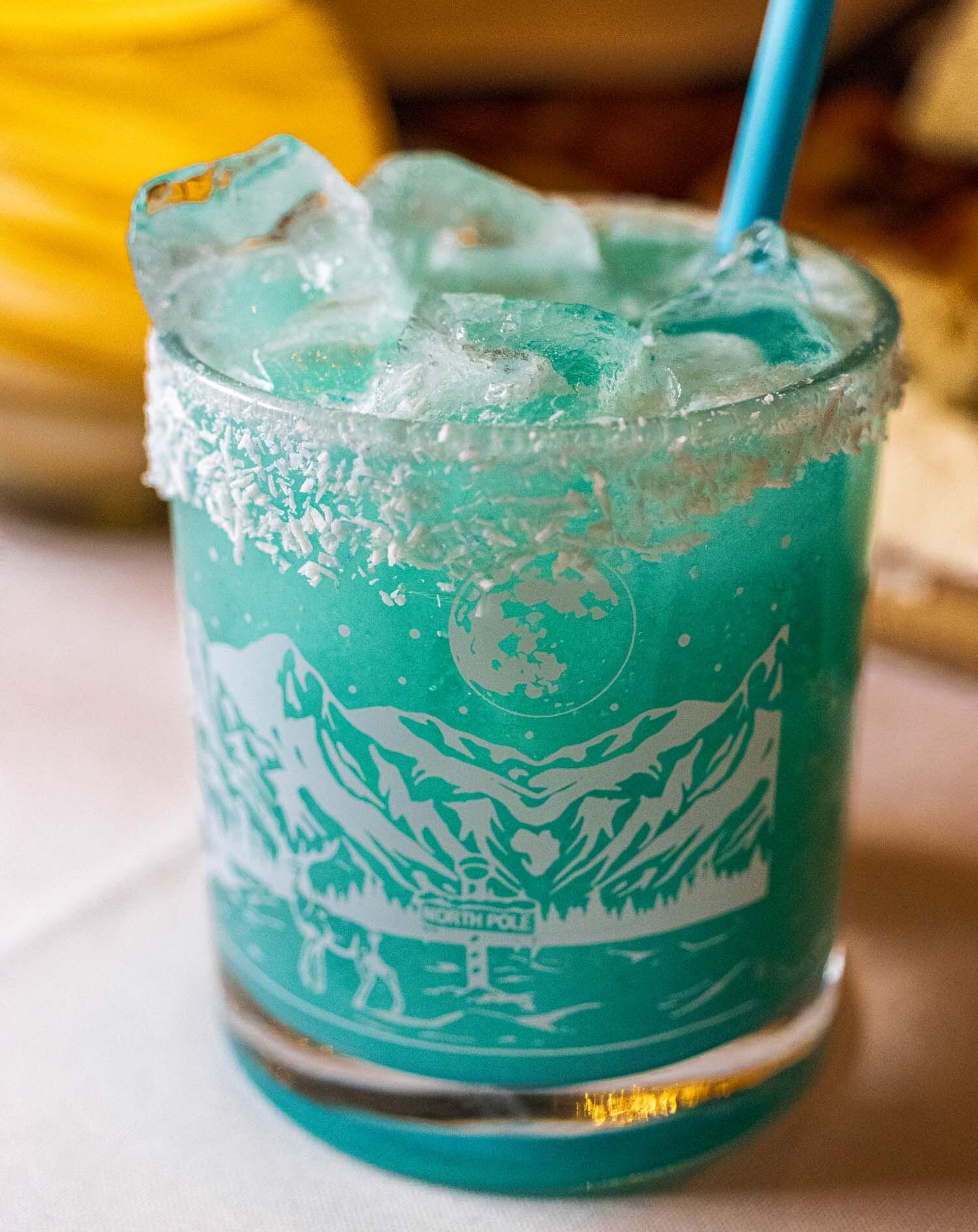 Tropical aqua-blue cocktail featuring Stoli Vanil, pineapple juice, cream of coconut, lime, and Triple Sec with a coconut rim and etched winter mountain scene glass design, served over crushed ice with a blue straw