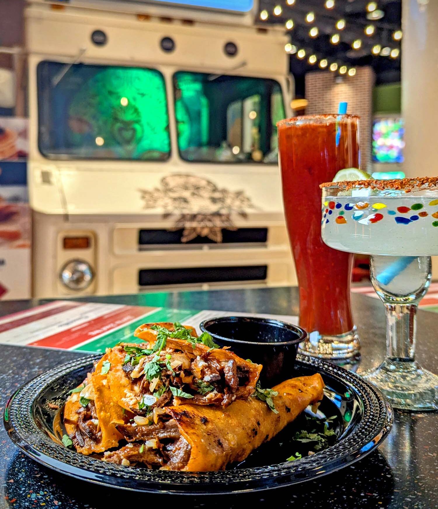 Birria tacos served in front of Distrito's vintage food truck display, accompanied by a tall red michelada and a festive margarita with colorful rim, string lights visible in background