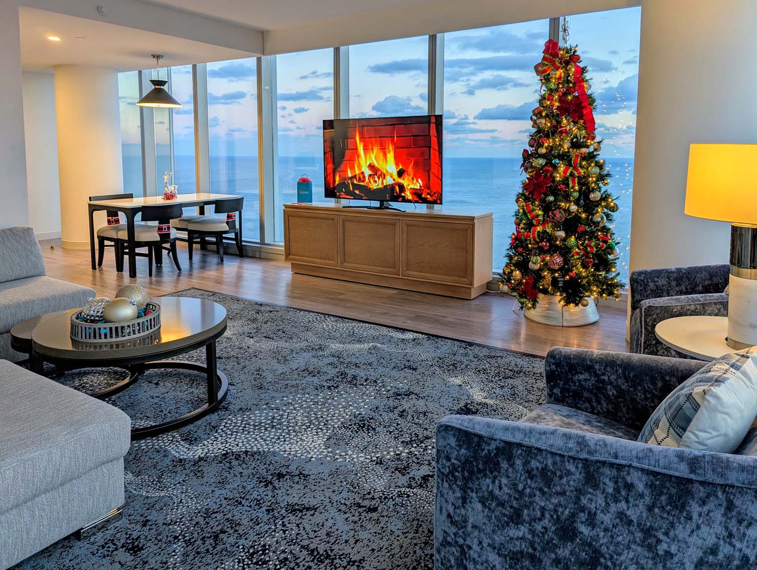 Luxury hotel suite living room featuring floor-to-ceiling windows overlooking the ocean, decorated Christmas tree, fireplace on TV screen, contemporary furniture, and modern decor