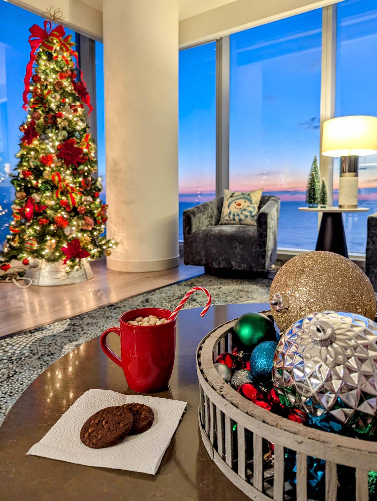 Red mug of hot chocolate with candy cane, chocolate cookies, and holiday ornaments with decorated Christmas tree and ocean view in background