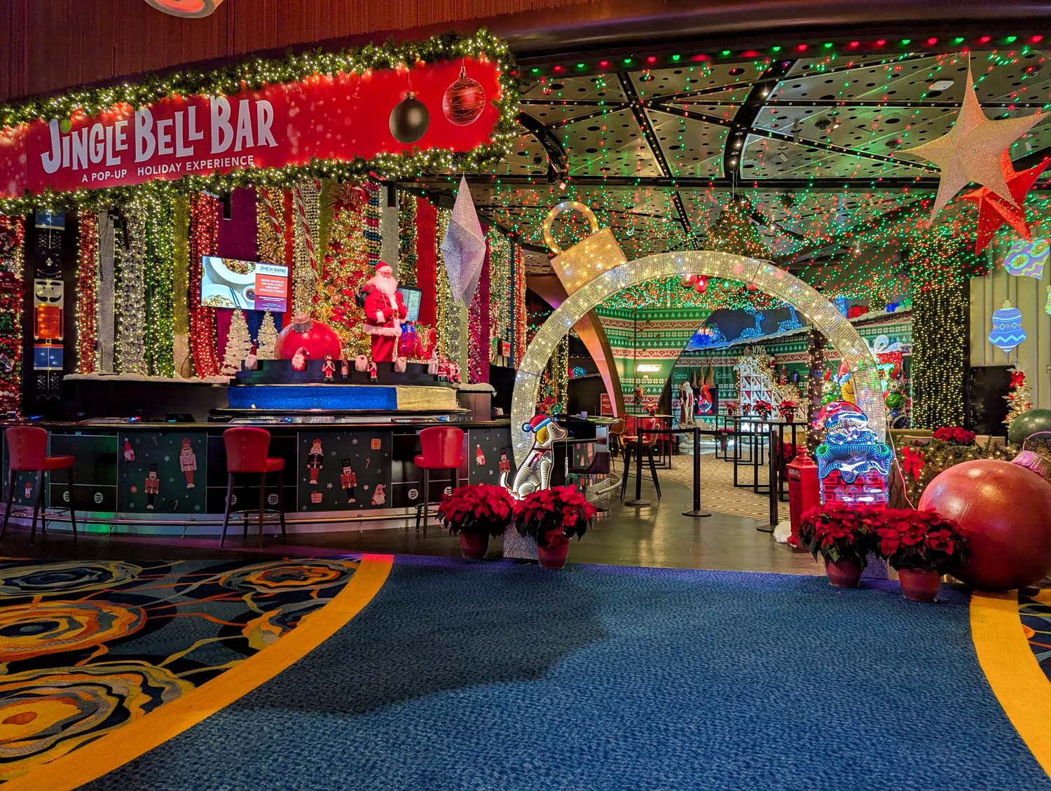 Elaborately decorated holiday themed bar with red and green lights, giant ornaments, glowing archway, Santa figure, poinsettias, and festive Christmas decor covering walls and ceiling