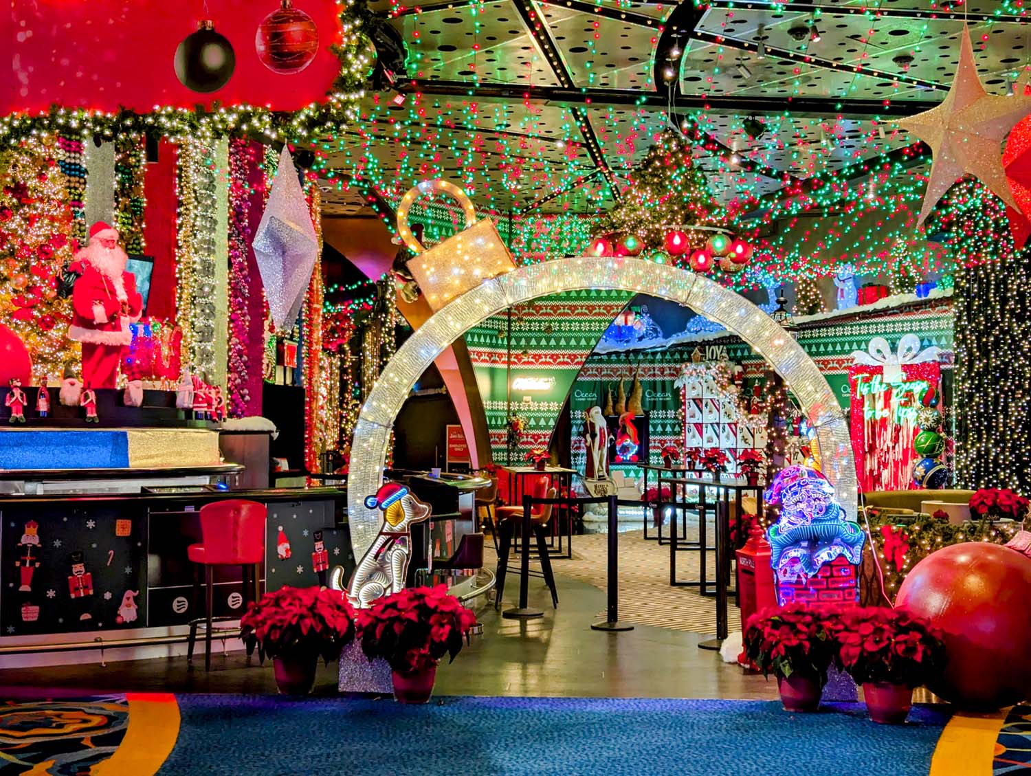 Immersive holiday-themed bar featuring a glittering archway entrance with LED light display, life-sized Santa Claus figure, red poinsettias in decorative pots, festive wall coverings with nutcracker patterns, ceiling decorated with thousands of red and green lights, and custom holiday-themed furniture and decor