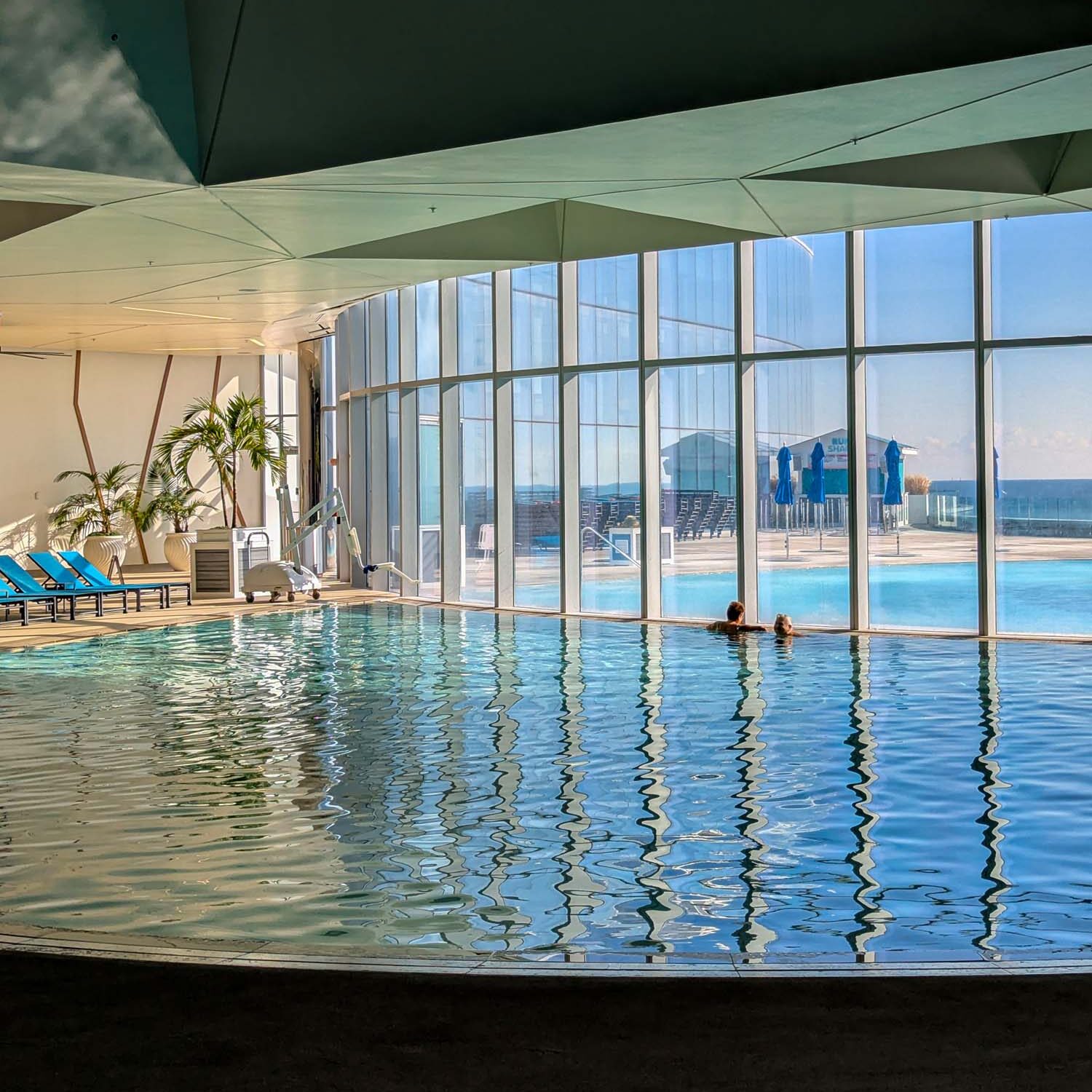 Ocean Casino Resort's indoor pool with floor-to-ceiling windows offering panoramic views of the Atlantic Ocean, featuring modern architectural elements, palm trees, and outdoor deck with blue lounge chairs