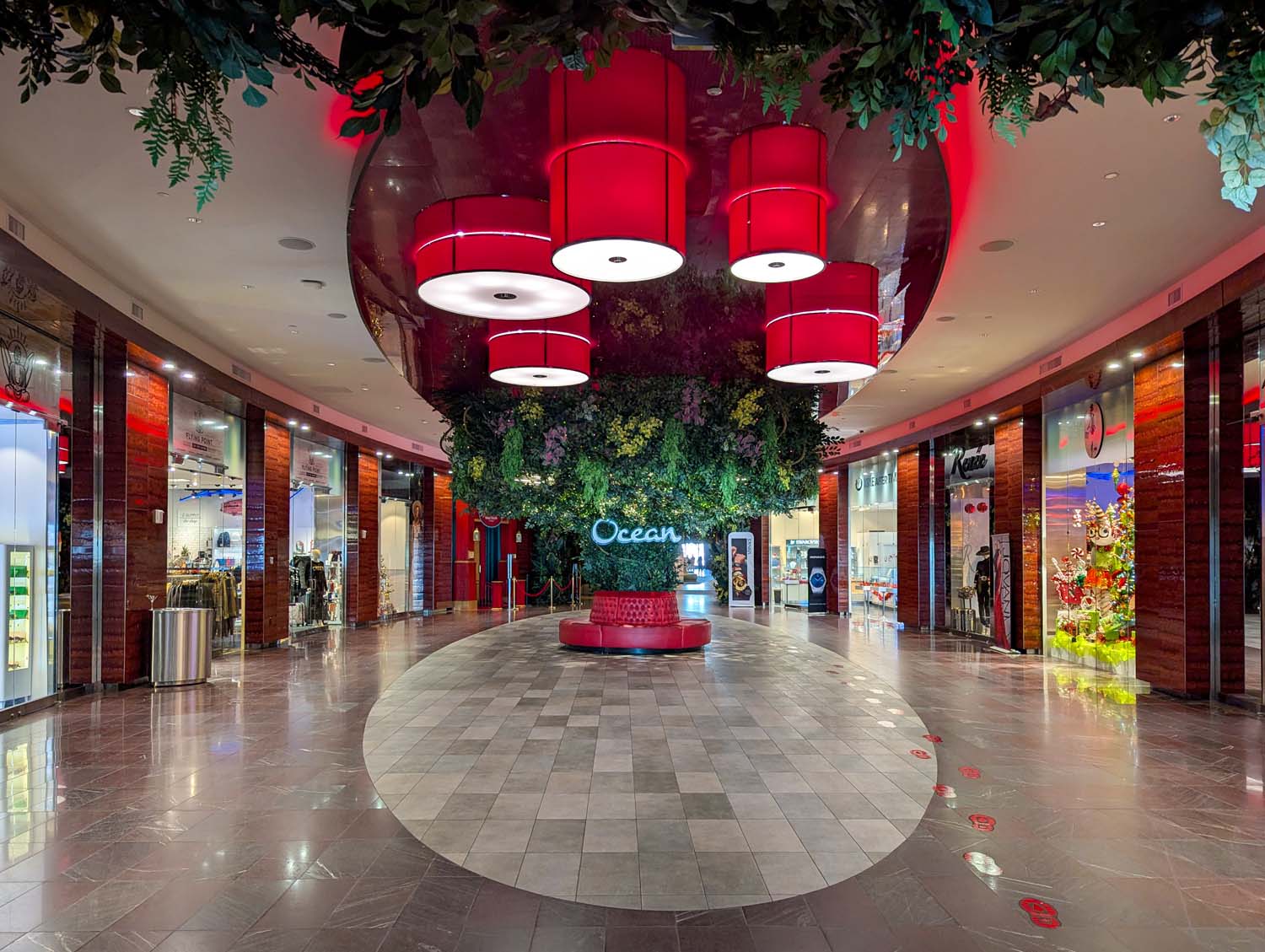 Luxury retail corridor at Ocean featuring red drum pendant lights, living wall installation with Ocean logo, marble floors, mahogany-colored walls, and holiday decorations reflecting in polished surfaces