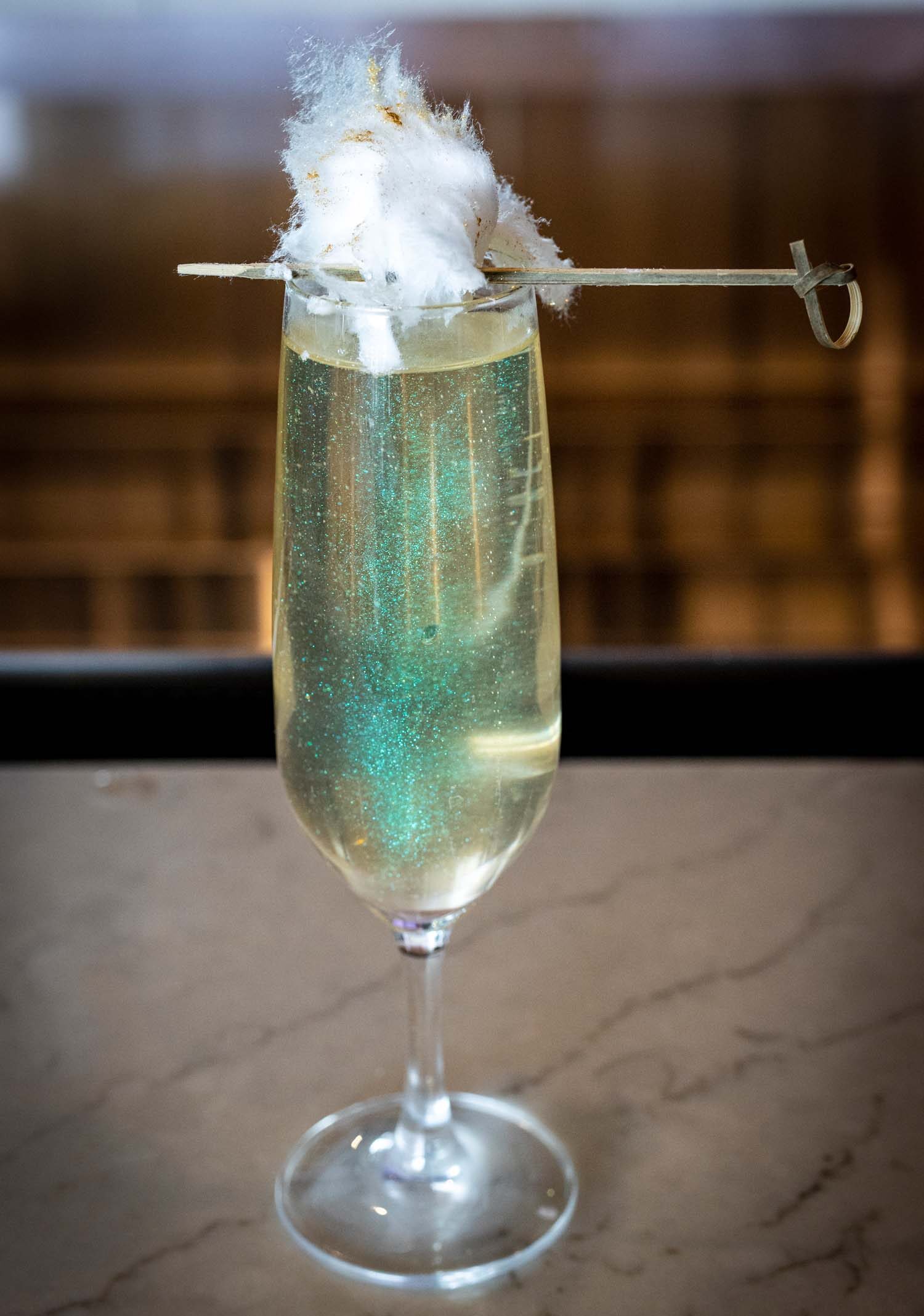 Champagne flute filled with sparkling prosecco and white cranberry juice, garnished with a cloud of cotton candy on top, featuring ethereal shimmer and served on a marble surface