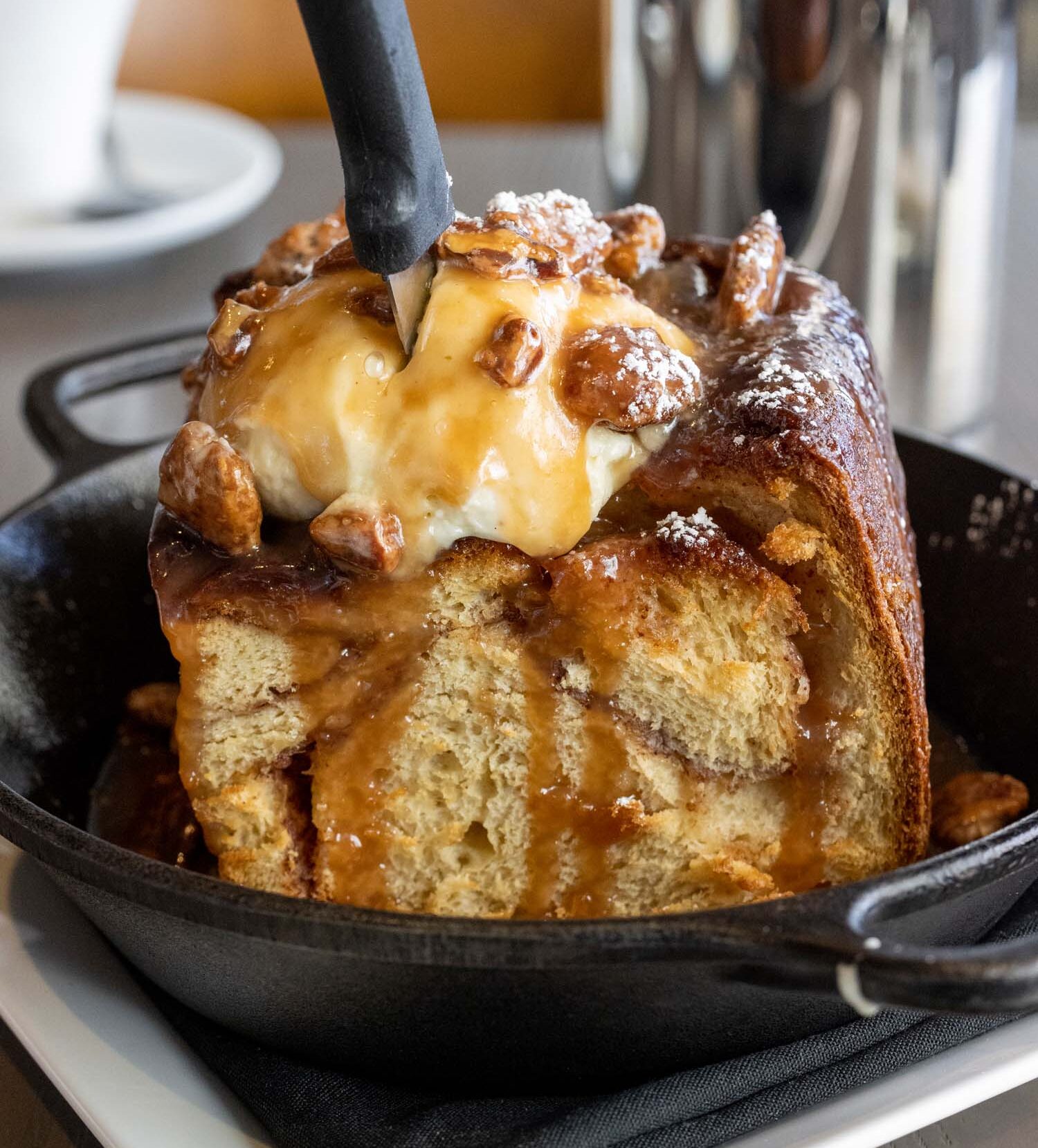 Close-up of a sticky bun in a cast iron skillet, topped with vanilla ice cream, candied pecans, and caramel sauce