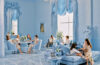 A luxurious blue and white room featuring ornate rococo details, with guests seated on upholstered chairs and chaises in pale blue damask. The space includes crystal chandeliers, elaborate wall moldings, and floor-to-ceiling windows with blue silk drapery. Fresh white floral arrangements accent the tables where guests are enjoying tea service