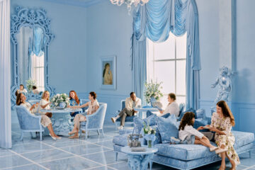 A luxurious blue and white room featuring ornate rococo details, with guests seated on upholstered chairs and chaises in pale blue damask. The space includes crystal chandeliers, elaborate wall moldings, and floor-to-ceiling windows with blue silk drapery. Fresh white floral arrangements accent the tables where guests are enjoying tea service