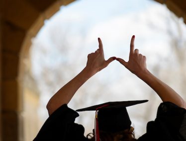 Graduate shows a W hand sign overhead while walking in silhouette through the arched portico of Bascom Hall