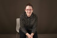 Rachel Willson-Broyles, dressed in dark colors, sitting on a white chair in a dark room, leaning toward the camera.