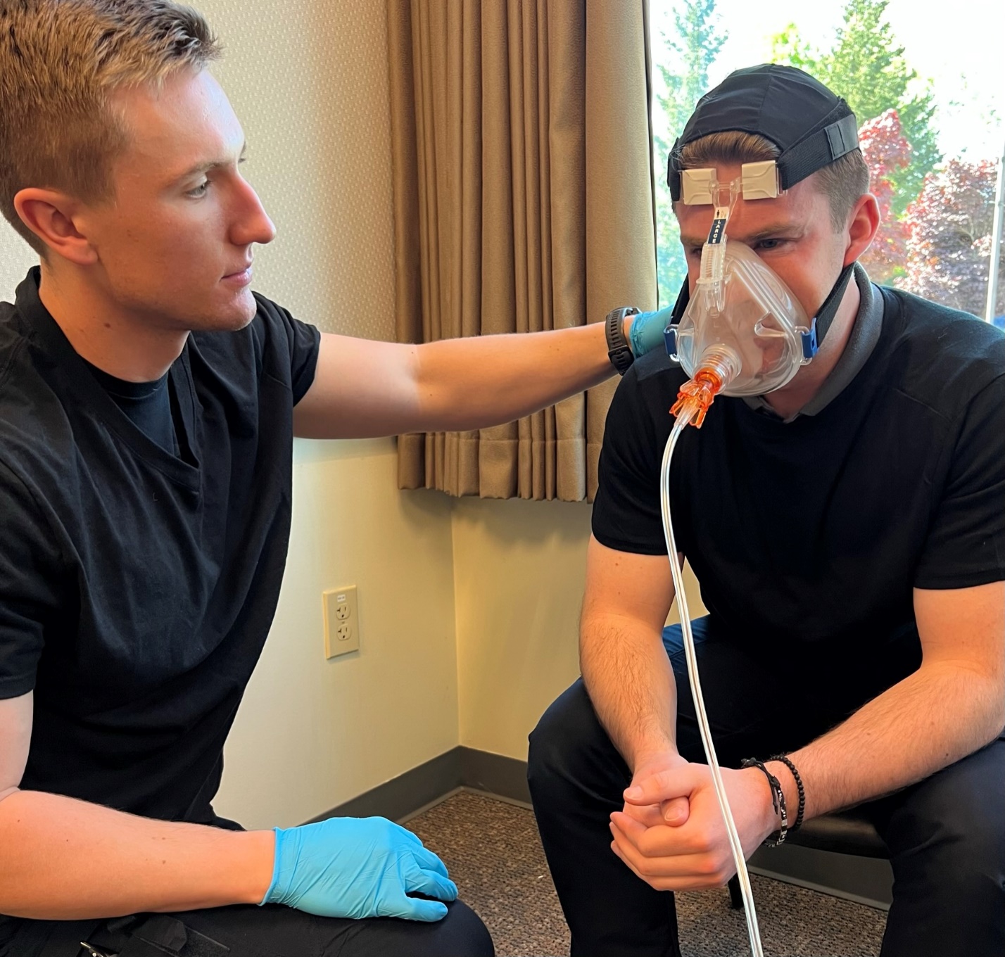 Gloved EMT kneeling next to seated patient and assuring/coaching after CPAP mask is secured.