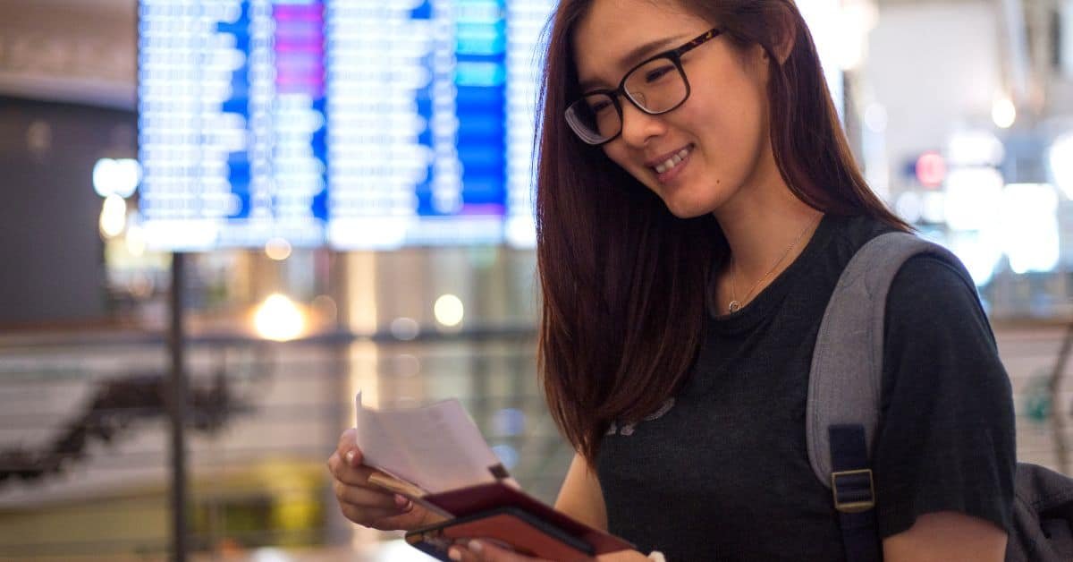 Young woman smiling while holding a passport and boarding pass at an airport, with flight information screens in the background.