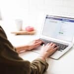 Person using a laptop to access Facebook, with a coffee cup and a pastry on the table, highlighting online social media engagement and remote work.