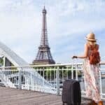 A traveler in a floral dress and sunhat stands on a bridge in Paris, gazing at the Eiffel Tower while holding a suitcase, symbolizing adventure and exploration.