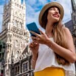 Young woman wearing a straw hat and casual outfit, smiling while holding a smartphone in front of a historic building. Bright blue sky and architectural details in the background. Ideal for travel and lifestyle content.