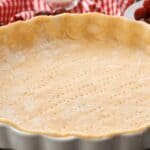 Unbaked pie crust in a fluted tart pan, set against a checkered red and white tablecloth, ready for filling with sweet or savory ingredients.