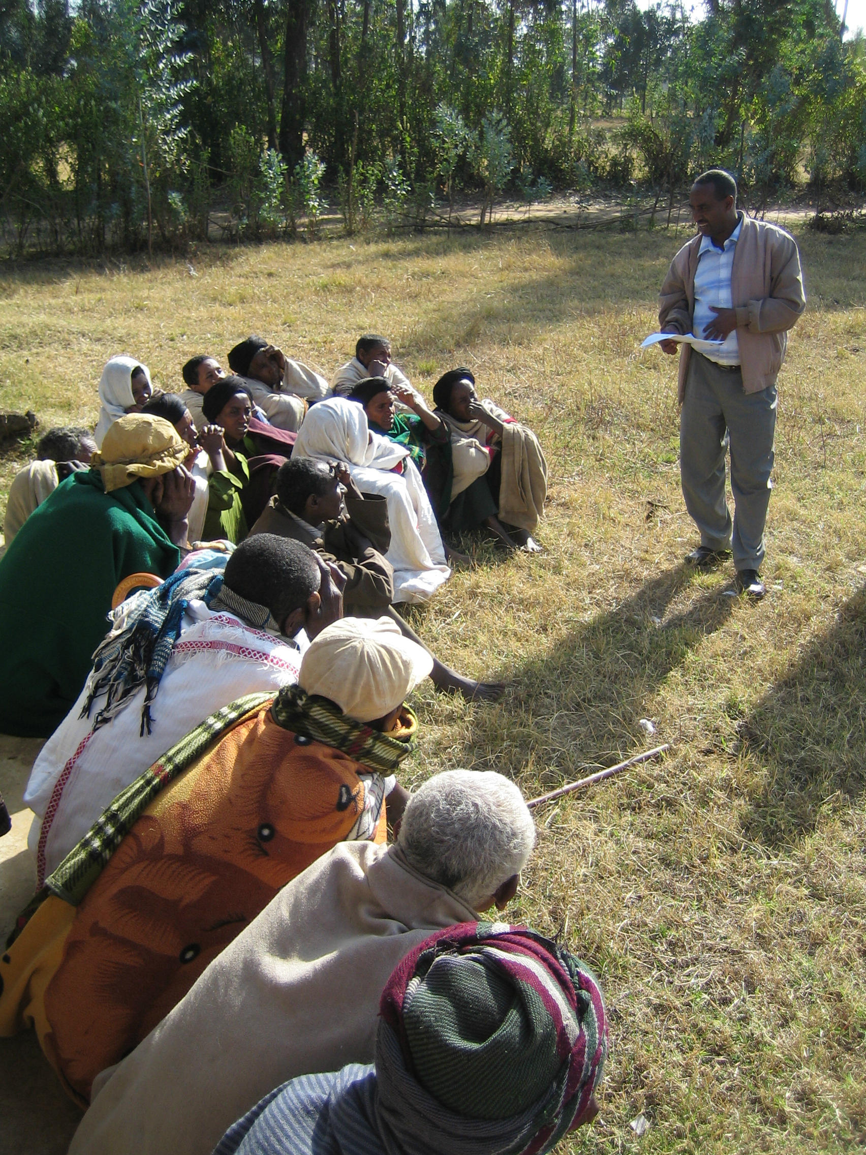 A standing speaker holds a paper and speaks to a group seated on grass outside on a sunny day, listening.