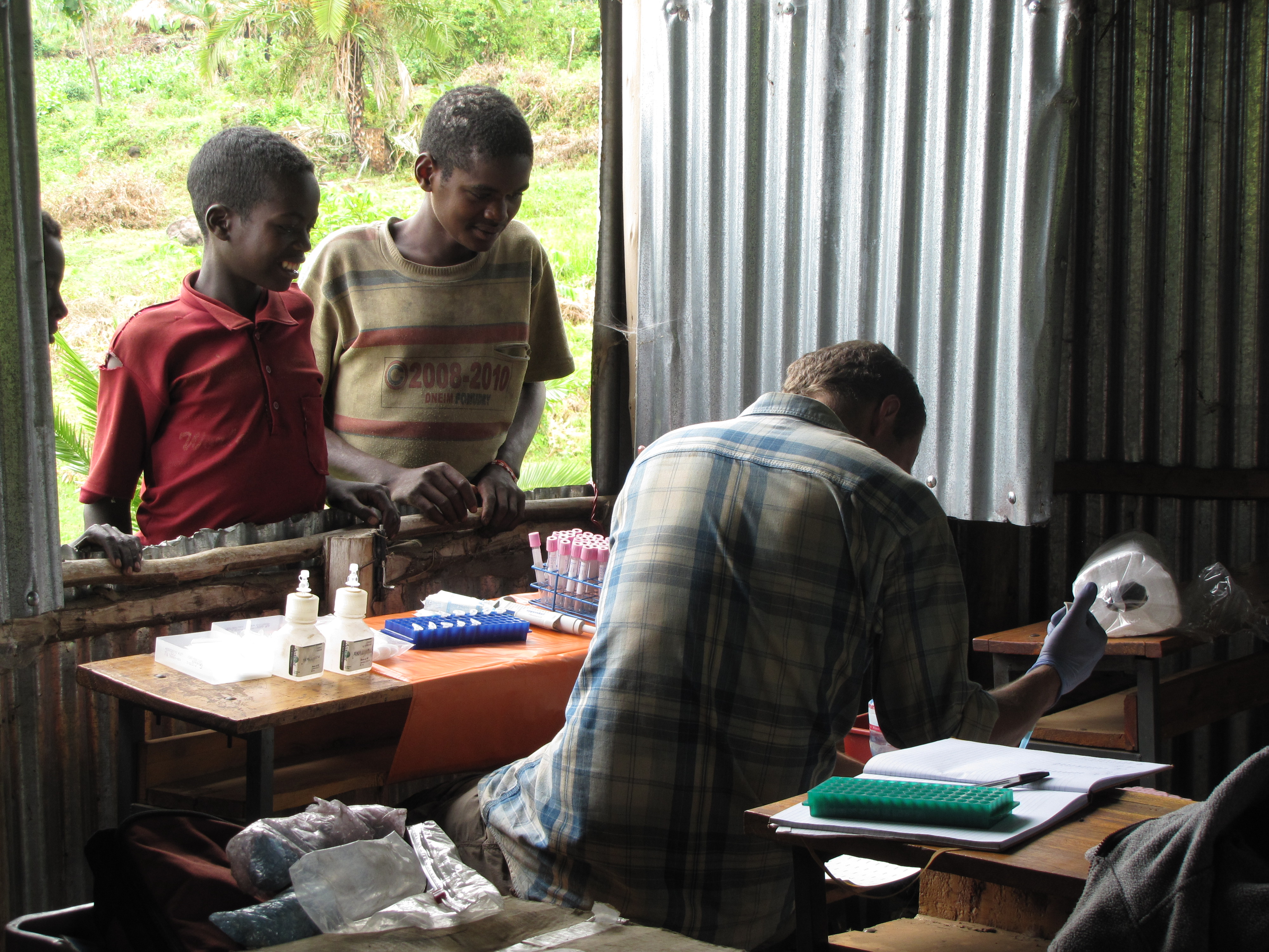 A person seated uses a pipette to place a sample in a test tube as two young people look on through a window.