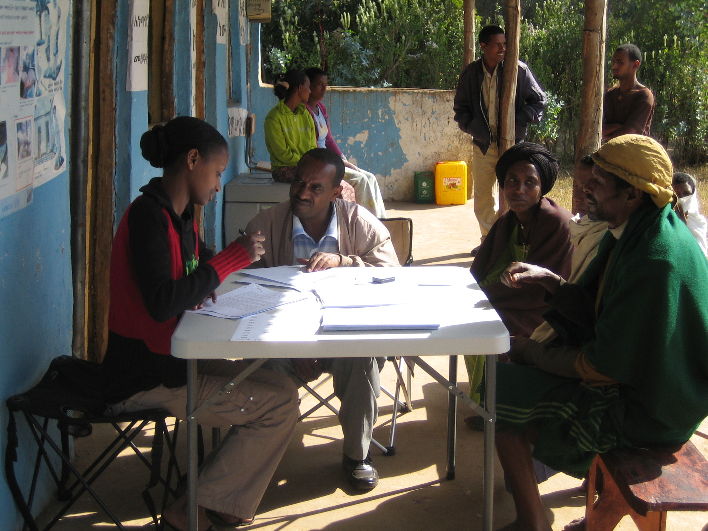 A person sits at a table collecting paperwork from a few other people who appear to be asking questions.