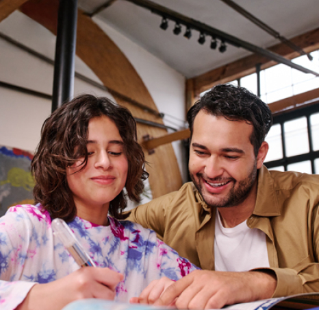 A father sits with his daughter at her desk, helping her with her work