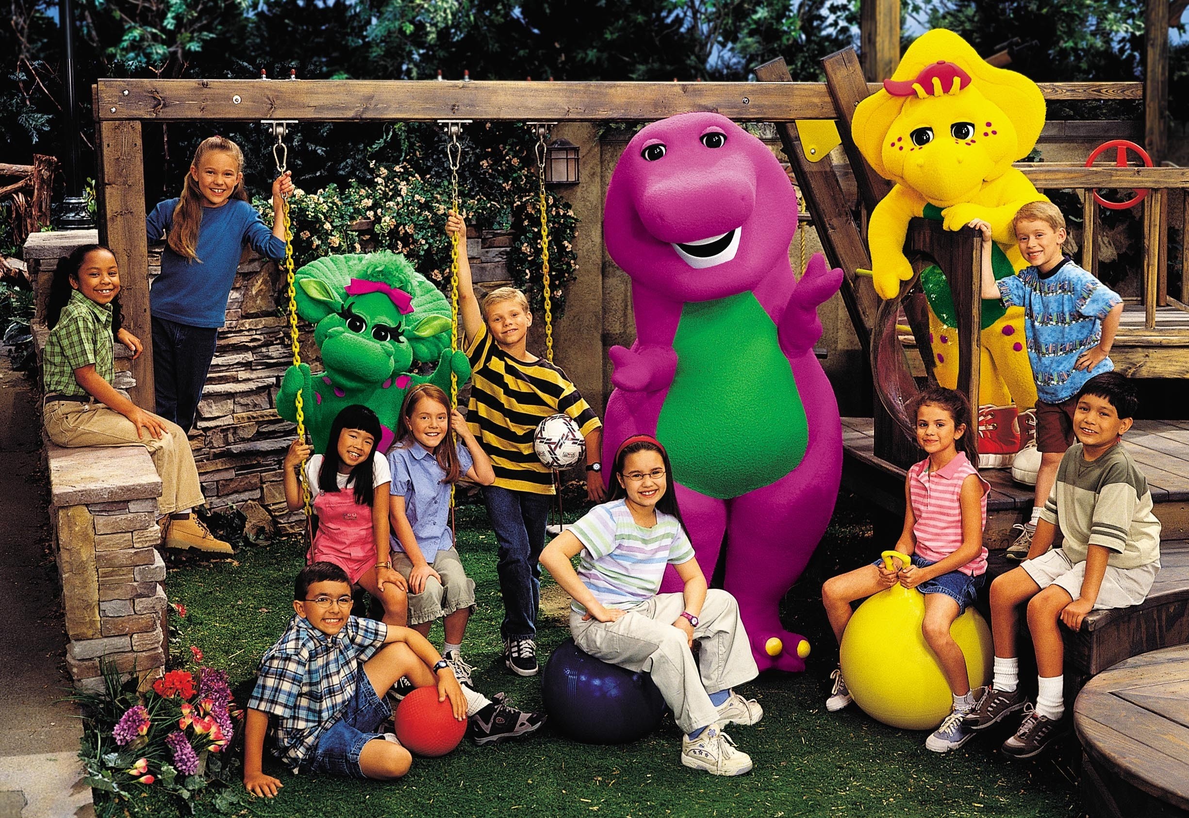 Foto do elenco do programa infantil Barney e Seus Amigos. Da esquerda para a direita na imagem: uma garota negra de olhos e cabelo escuros está sentada de lado em uma mureta, ela veste uma camiseta xadrez verde e calça e sapatos beges; uma garota branca de olhos e cabelo claros está apoiada com uma mão na corrente de um balanço, ela veste uma camiseta de manga longa azul e uma calça preta; um garoto branco de olhos e cabelo escuros está sentado no gramado com uma bola vermelha em uma de suas mãos, ele usa óculos de grau, veste uma camiseta xadrez azul, uma bermuda azul e calça um tênis preto com meias brancas; uma garota branca de olhos e cabelo escuros está sentada em um balanço ao lado de outra garota, ela veste uma camiseta branca coberta por uma peça rosa; a garota que divide o balanço é branca de olhos e cabelo claros, ela veste uma camiseta azul de botões, uma bermuda cinza e um tênis branco; atrás do balanço onde estão as duas meninas está Baby Bop, uma triceratops verde; um garoto branco de olhos e cabelo claros apoia uma mão na corrente do balanço enquanto segura uma bola de futebol com a outra, ele veste uma camiseta listrada preta e amarela, uma calça jeans azul e um tênis preto e branco; Demi Lovato, pele branca de olhos e cabelos escuros está sentade em uma bola azul, elu usa óculos de grau, veste uma camiseta listrada branca e azul, uma calça e tênis brancos; Barney, um tiranossauro rex magenta está ao lado de Lovato; Selena Gomez, uma garota branca de olhos e cabelo escuros está sentada em uma bola amarela, ela veste uma regata listrada vermelha e branca, uma bermuda jeans, um tênis cinza e meias brancas; B.J, um protoceratops amarelo está atrás de Gomez; um garoto branco de olhos e cabelo escuros está sentado na escada, ele veste uma camiseta de manga longa cinza, uma bermuda branca, um tênis vinho e meias brancas; o último garoto da direita está em pé na escada, ele é branco de olhos e cabelo claros, ele veste uma camiseta azul, uma bermuda marrom, um tênis cinza e meias brancas. O ambiente é um parque de tom marrom e gramado verde