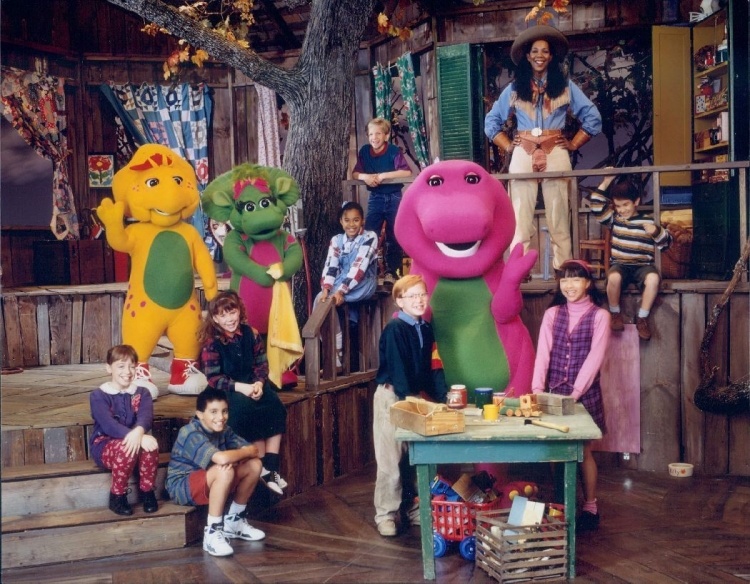 Foto do elenco do programa infantil Barney e Seus Amigos. Da esquerda para a direita na imagem: uma garota branca de olhos e cabelo claros está sentada na escada, ela veste um casaco azul, uma calça vermelha e um sapato preto; B.J, um protoceratops amarelo, está em pé; um garoto branco de olhos e cabelo escuros está sentado na escada, ele veste uma camiseta cinza, uma bermuda vermelha, um tênis branco e meias brancas; uma garota branca de olhos e cabelo claros está sentada na escada, ela veste uma camiseta de manga longa listrada vermelha, uma peça preta, um tênis branco e meias pretas; Baby Bop, uma triceratops verde está em pé enquanto carrega um cobertor amarelo; uma garota negra de olhos e cabelo escuros está sentada no apoio da escada, ela veste uma camiseta de manga longa branca quadriculada com uma peça jeans; um garoto branco de olhos e cabelo claros está apoiado no apoio da escada, ele veste uma camiseta azul e vermelha e uma calça jeans; um garoto branco de olhos e cabelo claros está em pé, ele usa um óculos de grau, veste uma camiseta de manga longa azul, uma calça bege e um sapato bege; Barney, um tiranossauro rex magenta está em pé; uma garota branca de olhos e cabelo escuros está em pé, ela veste uma camiseta de manga longa rosa com uma peça de botões roxa; uma mulher negra de olhos e cabelos escuros está em pé, ela veste um figurino clássico de cowboy nos tons azul e bege; o último garoto da direita está sentado, ele veste uma camiseta de manga longa listrada branca e marrom, uma bermuda marrom, um tênis marrom e meias brancas. O ambiente é uma oficina de madeira no tom marrom.