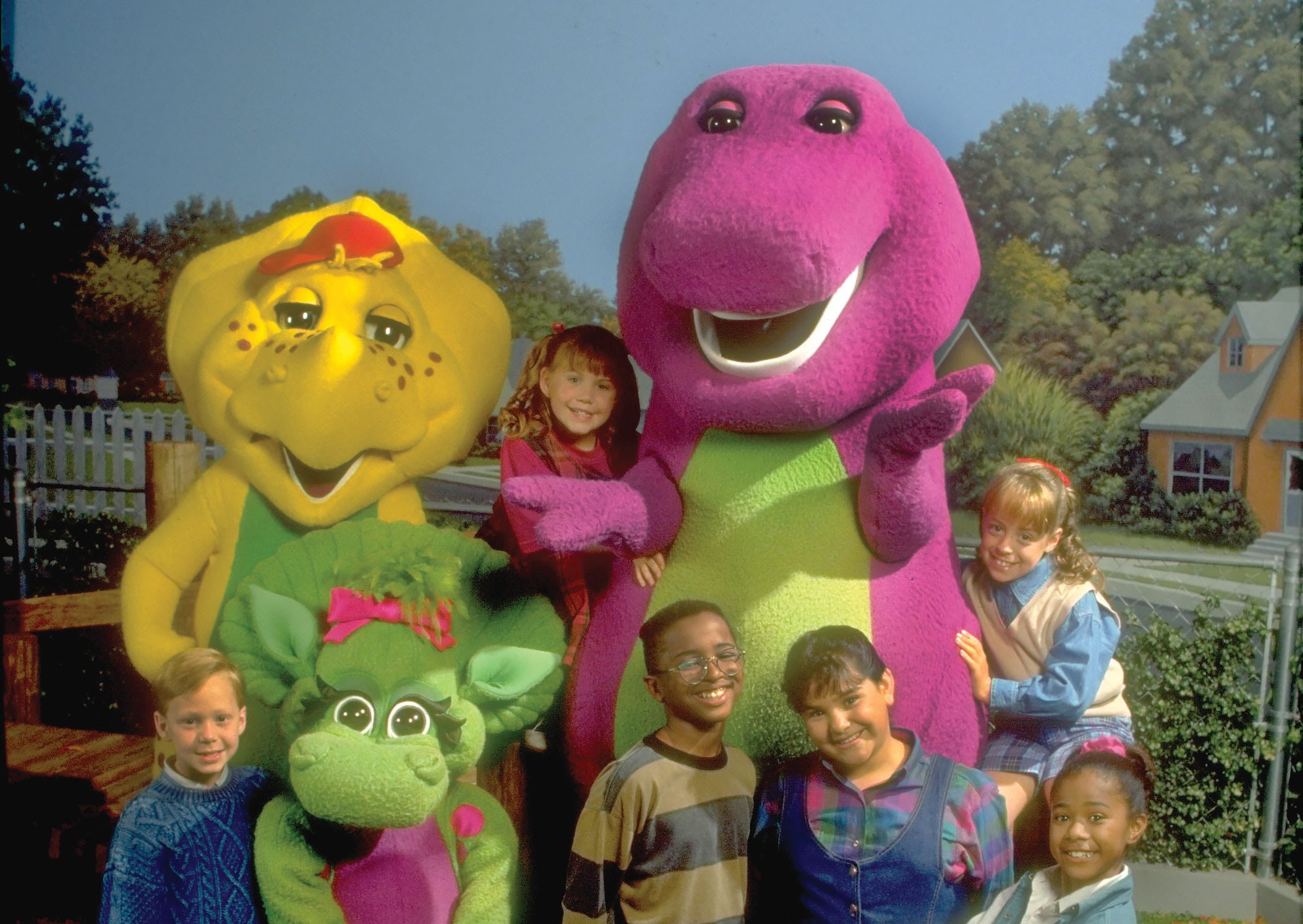 Foto do elenco do programa infantil Barney e Seus Amigos. Da esquerda para a direita na imagem: um garoto branco de olhos e cabelo claros, ele veste um casaco azul; B.J, um protoceratops amarelo; Baby Bop, uma triceratops verde; uma garota branca de olhos e cabelo claros, ela veste rosa; Barney, um tiranossauro rex magenta; um garoto negro de olhos e cabelo claros, ele usa um óculos de grau e veste uma camiseta de manga longa listrada preta e cinza; uma garota branca de olhos e cabelos escuros, ela veste uma camiseta de botões e uma peça azul; uma garota negra de olhos e cabelo escuros, ela veste uma jaqueta branca e azul; uma garota branca de olhos e cabelo claros, ela veste uma camiseta de manga longa azul com uma peça bege e uma bermuda listrada azul. O ambiente é um bairro bem arborizado. Todos estão sorrindo para a câmera.