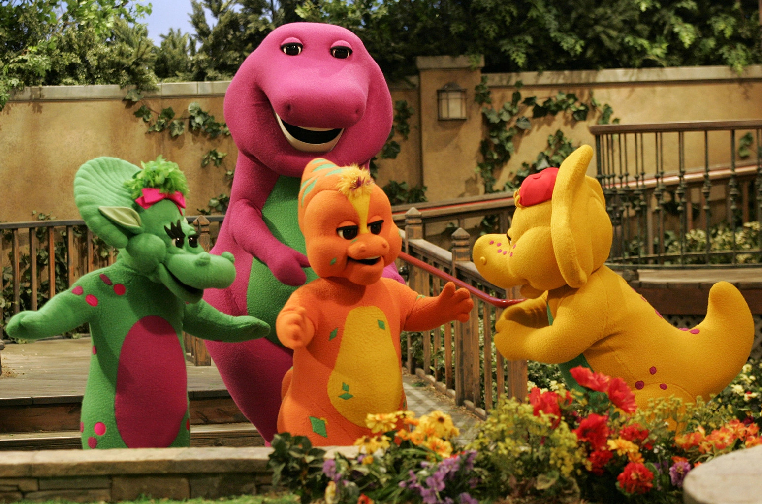 Foto do elenco do programa infantil Barney e Seus Amigos. Da esquerda para a direita na imagem: Baby Bop, uma triceratops verde; Barney, um tiranossauro rex magenta; Riff, um hadrossauro laranja; B.J, um protoceratops amarelo. Todos estão se divertindo e sorrindo. O ambiente é repleto de escadas de madeira no tom marrom. Há muitas árvores, plantas e flores coloridas ao redor.