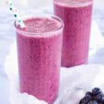 Blueberry pineapple smoothie in two glasses with a straw against a grey background.