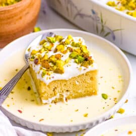 Rasmalai tres leches cake served on a white plate, topped with pistachios and the sweetened milk mixture.