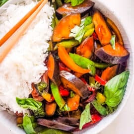 Thai basil eggplant in a white bowl, served with rice and chopsticks on the side.