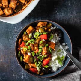 Vegan kung pao tofu in a dark blue bowl with white rice and sprinkled with green onions next to a grey towel, with a fork in the bowl.