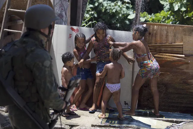 Children play in an outdoor shower as a soldier takes part in a surprise operation in the Manguinhos slum in Rio de Janeiro, Brazil, Thursday, January 18, 2018. (Photo by Leo Correa/AP Photo)