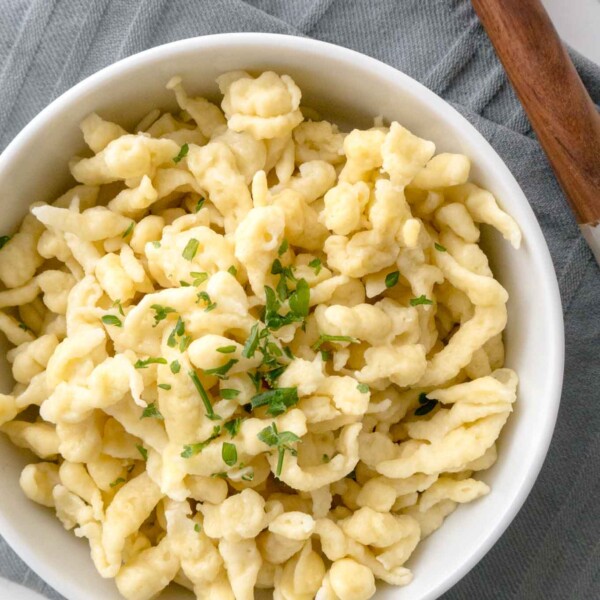 Top-down shot of a white bowl of spaetzle garnished with parsley on a grey dishtowel next to a wooden spoon.