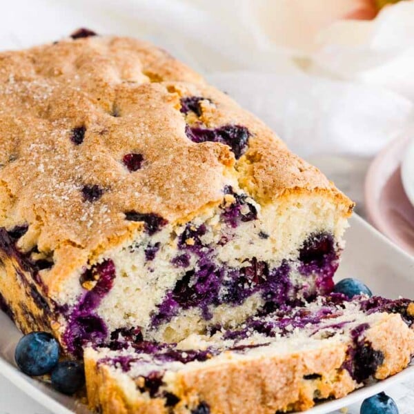 A loaf of blueberry bread on a white, rectangular plate with 1 slice cut off lying in front of it, garnished with blueberries and a flower.