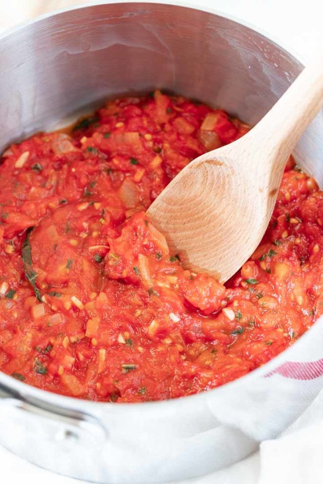 A stainless steel pot with tomato sauce, with a wooden cooking spoon in it on a white dish towel.