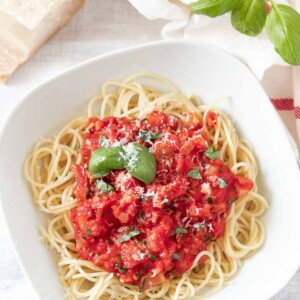 A white plate, with spaghetti topped with tomato sauce, garnished with basil leaves and parmigiano. Next to it, there's basil leaves, a white dish towel with a red stripe and a block of Parmigiano cheese.