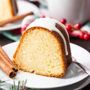 A piece of bundt cake with white glazing on top on a white plate with a fork and two sticks of cinnamon. There are cranberries, a cup and another plate in the background