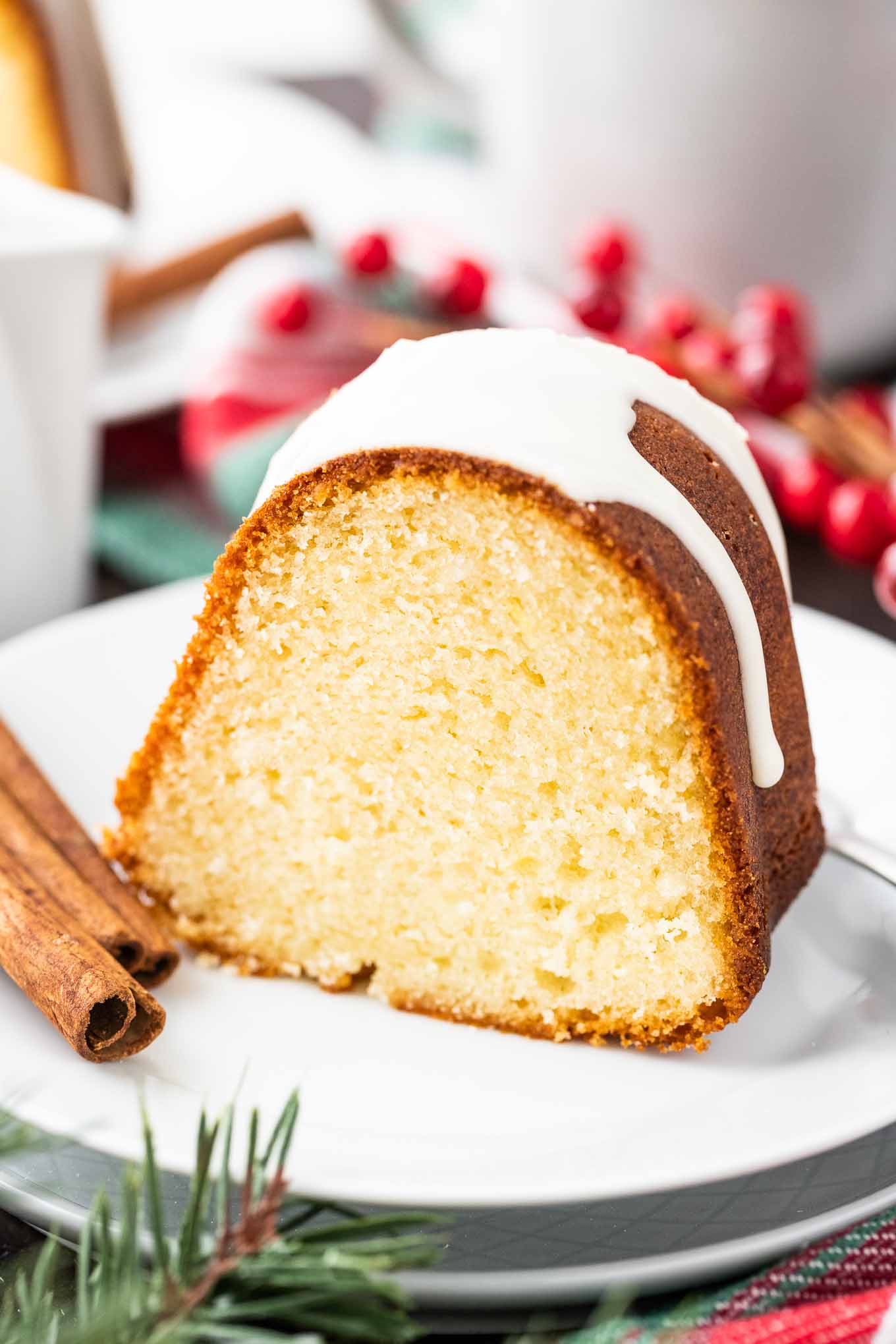 Close-up of a piece of bundt cake with white glazing on top on a white plate with a fork and two sticks of cinnamon. There are cranberries, a cup and another plate in the background.