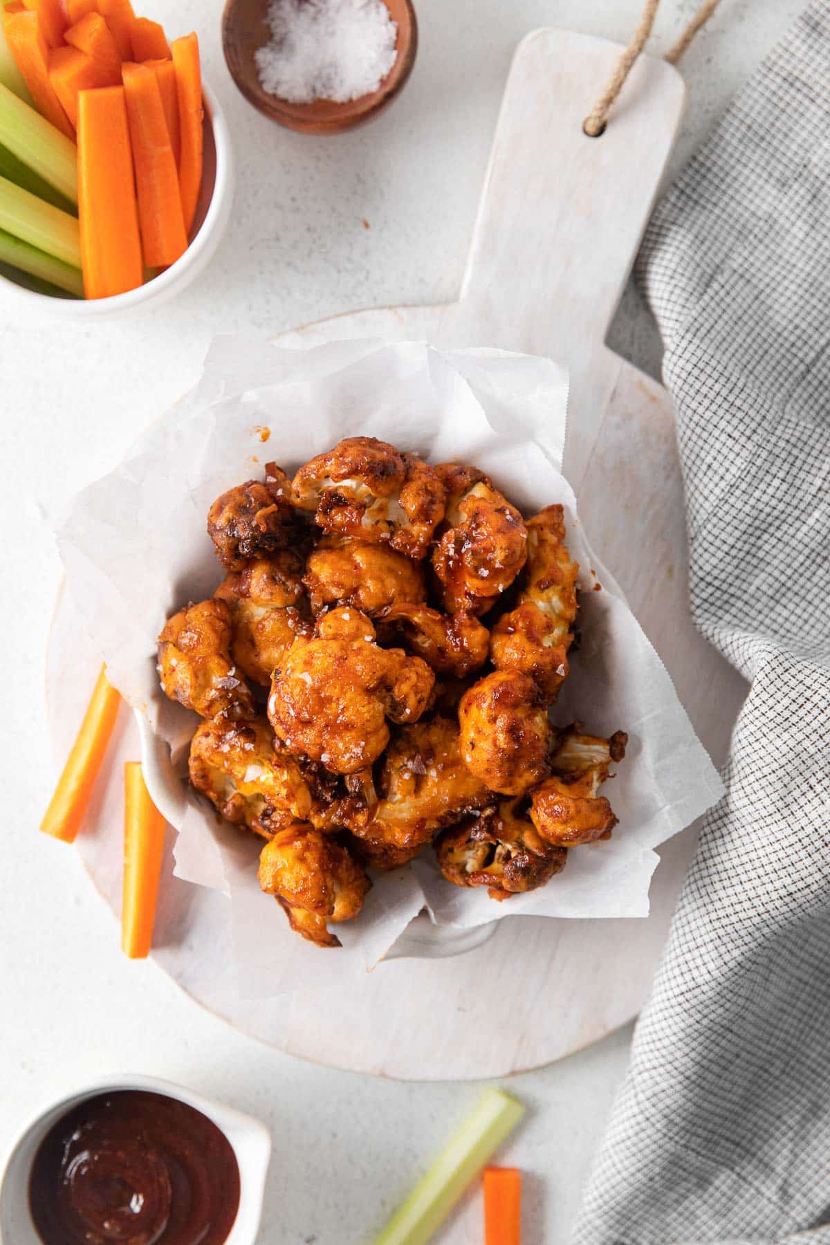 Cauliflower wings in a serving dish on a wooden board, next to it are some sliced vegetables in a small bowl and some BBQ sauce.