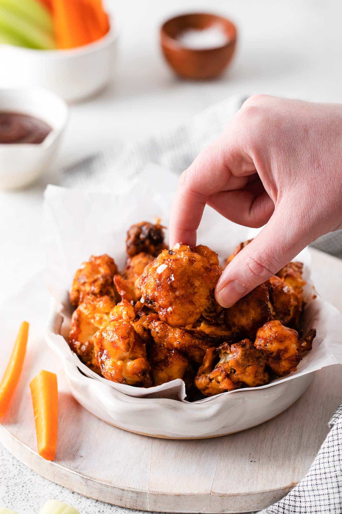 A female hand holding a cauliflower wing coated in BBQ sauce next to a white bowl filled with more wings.