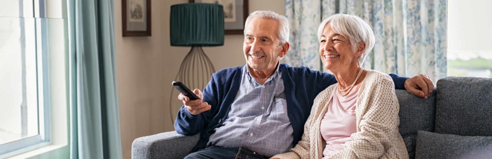 An elderly couple is sitting together on a grey couch, watching television. The man on the left is holding a remote control and wearing a blue cardigan over a light blue shirt. The woman on the right is dressed in a light pink top with a beige knitted cardigan, and they both appear to be smiling. Behind them is a green lamp and a pair of framed pictures on the wall. Light blue curtains with a floral pattern are drawn to the sides of a large window that lets in natural light.