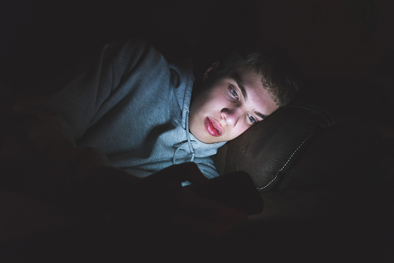 A young person lies comfortably, illuminated by their phone's light, deeply focused. This scene reflects how technology influences youth engagement in discussions about policing and social issues.