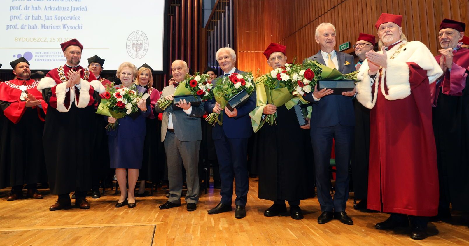 Laureates of the Laurus Aesculapii award on stage along with prof. Andrzej Tretyn, NCU Rector and prof. Dariusz Grzanka, Vice-Rector for Collegium Medicum. Laureates are holding bouquets of flower and awards.