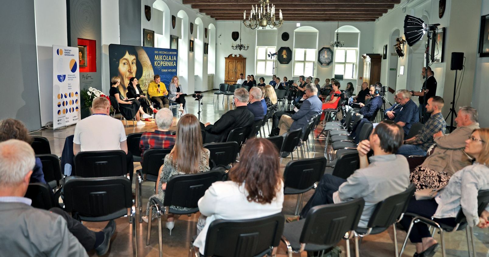 Debata organizowana z okazji 550. rocznicy urodzin Mikołaja Kopernika, w której uczestniczy czworo ludzi. Widać także widownię na krzesłach. Debata odbywa się w sali mieszczańskiej Muzeum Okręgowego