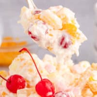 image of a spoon full of ambrosia salad with a cup full of ambrosia salad with cherries on top. On top of the image is is the title of the post with green and black lettering