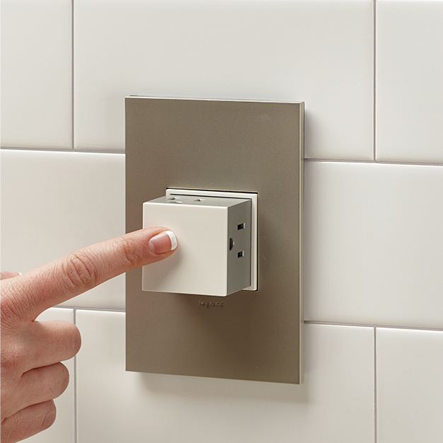 Finger placed on an adorne pop-out outlet with bronze wallplate on a white tile backsplash