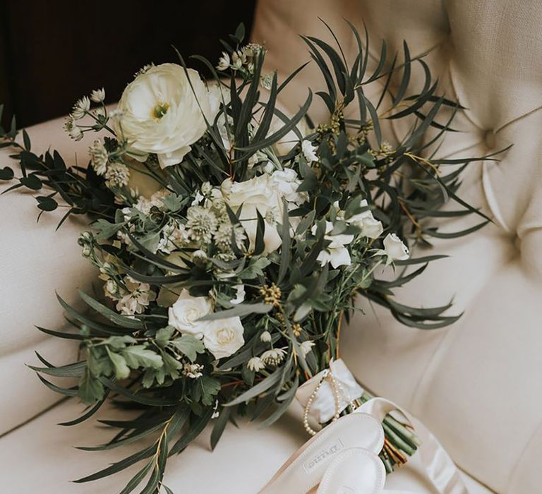 Simple wedding bouquet with white flowers and foliage tied with white ribbon and white satin heeled shoes 